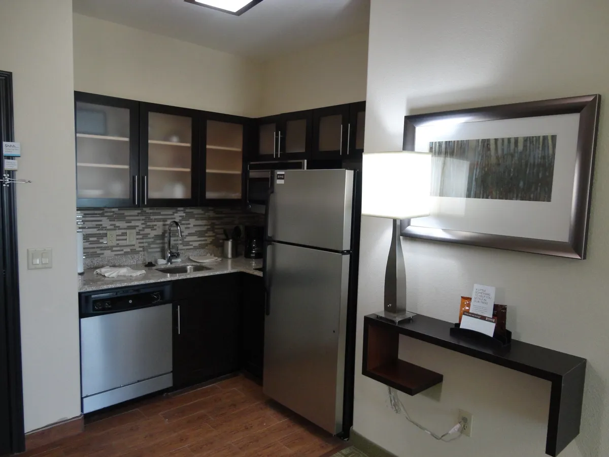 A contemporary kitchen featuring a stainless steel refrigerator, modern cabinetry, a textured countertop, decorative artwork, and a stylish lamp. The space is well-lit and inviting, designed to support both everyday cooking and entertaining guests.