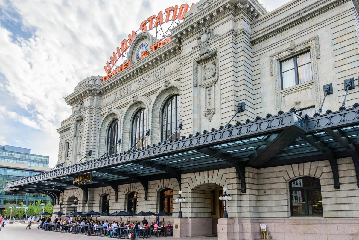 The image captures Denver’s iconic Union Station, with its recognizable sign, grand arched windows, and bustling outdoor seating area surrounded by intricate architectural details. People are dining at outdoor cafes, taking photos, and travelers are coming and going, creating a lively urban atmosphere.