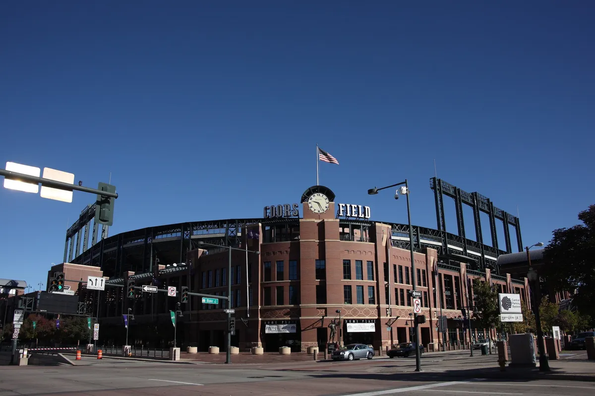 This exterior image captures the vibrant Coors Field Stadium with its iconic clock tower and American flag, surrounded by the lively atmosphere of the Ballpark neighborhood. You can see people gathering for a baseball game, as well as nearby dining options and festive street life.