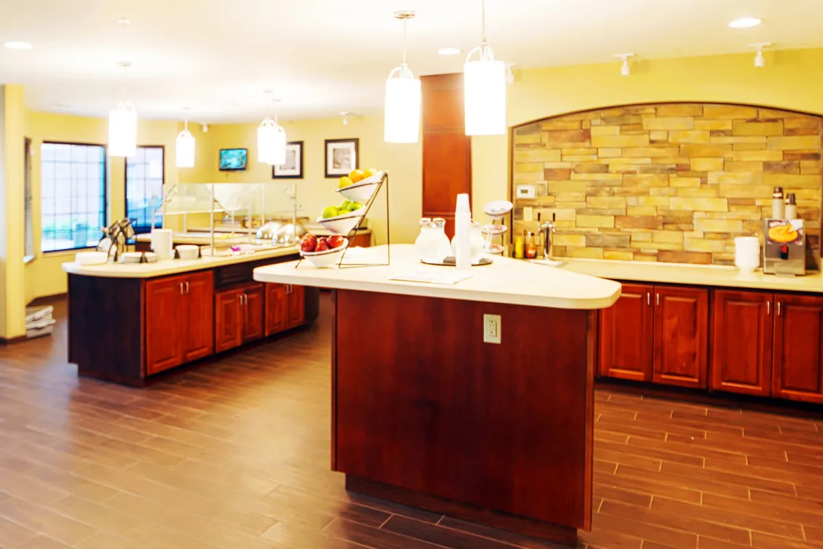 An inviting dining area centered around a large island counter with a vibrant fruit display, stone wall accents, and hanging light fixtures. The space feels communal and is set up for both casual breakfasts and gatherings with family or friends.