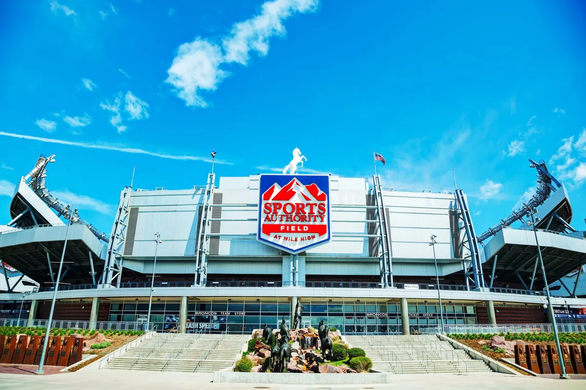 This exterior shot showcases the impressive stadium architecture of Sports Authority Field, with the bold signage and an expressive sculpture in the foreground. The energy of fans gathering for live sports or community events is palpable, capturing the pulse of Denver.