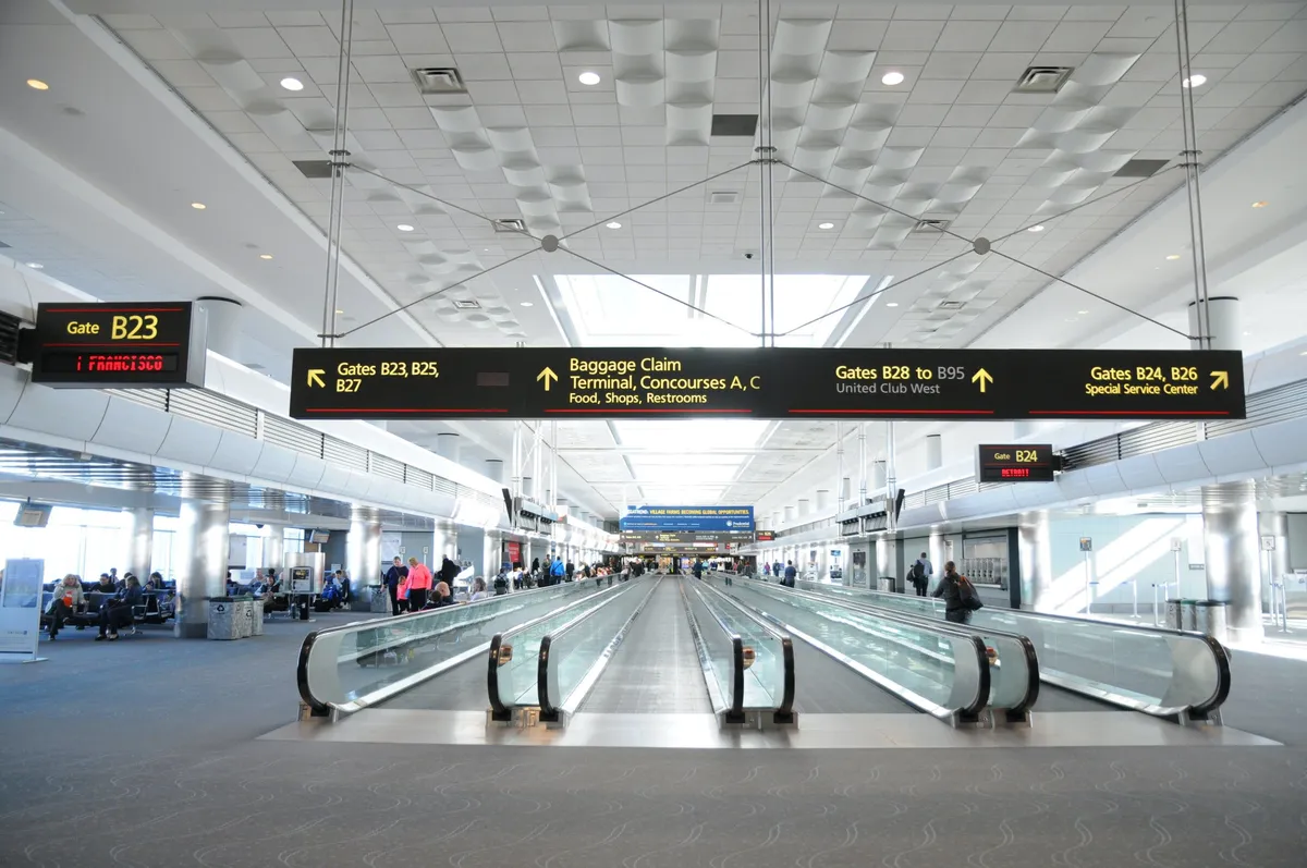 A busy interior scene at an airport terminal with moving walkways, clear gate signage, numerous seating areas, and passengers in transit. The image conveys the movement and connectivity of business travelers arriving or departing from Denver.