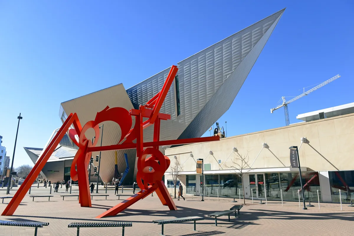 A striking large red sculpture anchors the scene in front of a modern building, with people walking and engaging in casual gatherings. The urban energy and contemporary architecture highlight Denver’s thriving downtown environment and creative spirit.