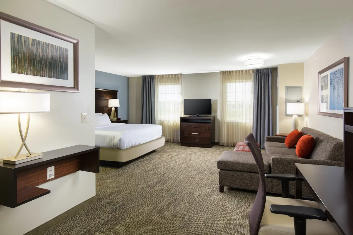 The image showcases a bright, comfortable suite interior with a plush bed, inviting sofa, flat-screen television, large windows letting in natural light, and a warm lamp, creating a cozy atmosphere ideal for relaxing or unwinding after a day in Denver.