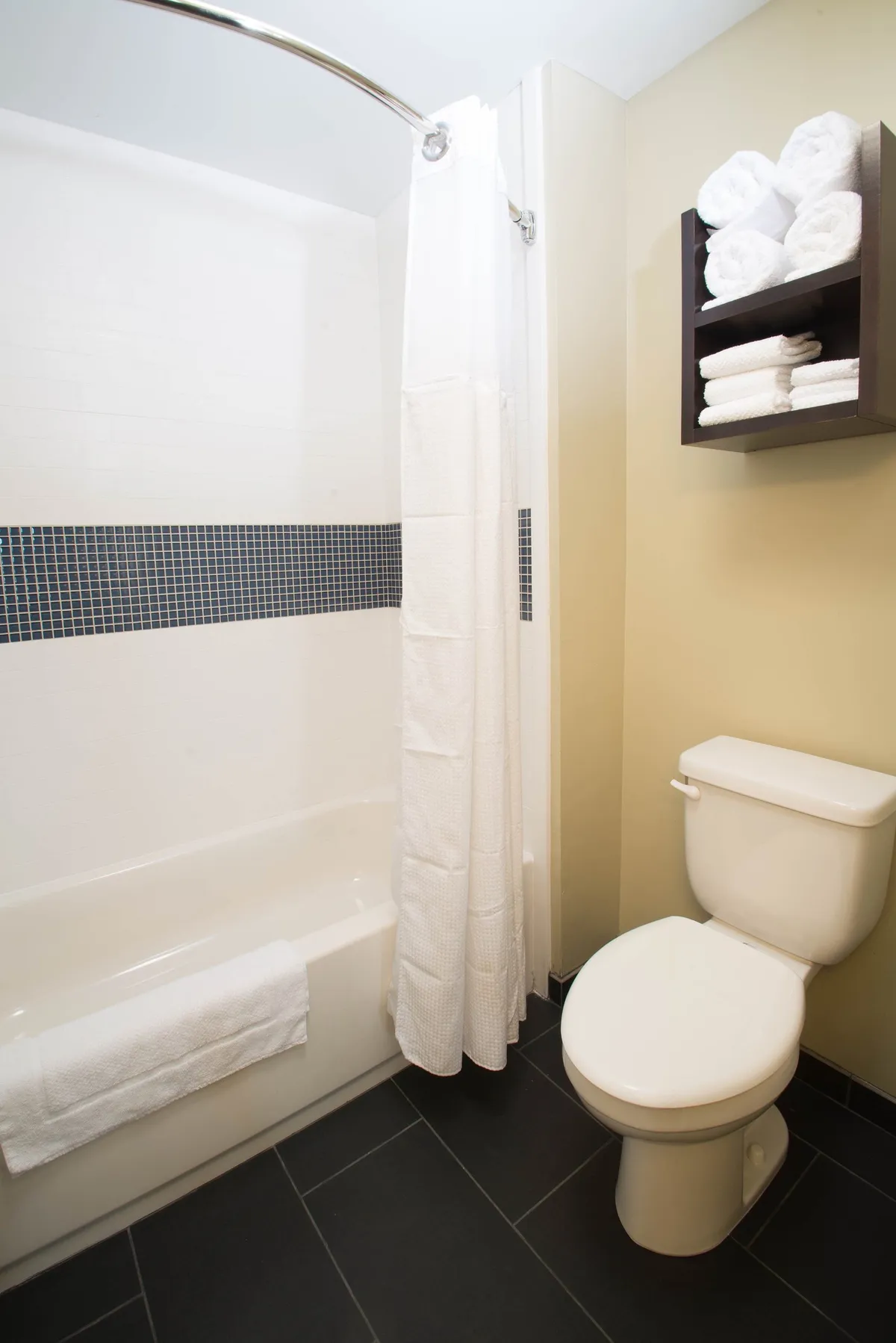 A bright, spa-inspired bathroom featuring a bathtub with a shower curtain, a towel rack stacked with plush towels, and a toilet. The clean lines, soft lighting, and calming decor evoke a sense of refreshment and relaxation, perfectly aligning with the idea of starting or ending your day in comfort.