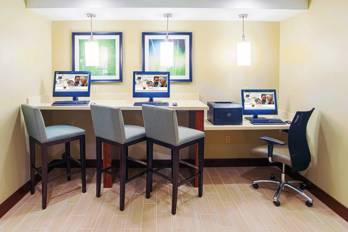 A modern workspace area equipped with computer workstations, stylish hanging light fixtures, and framed artwork. The environment is bright and conducive to productivity, offering a contemporary setting for work or leisure activities.