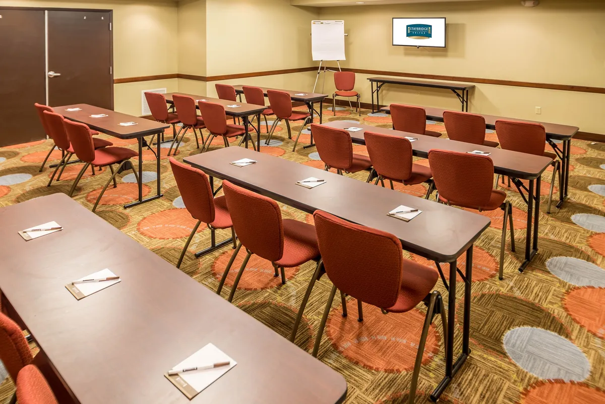 A spacious hotel meeting room with neatly arranged rows of tables and bold red chairs, facing a large presentation screen. The environment is bright and conducive to focused collaboration, hinting at team meetings, workshops, and social gatherings.